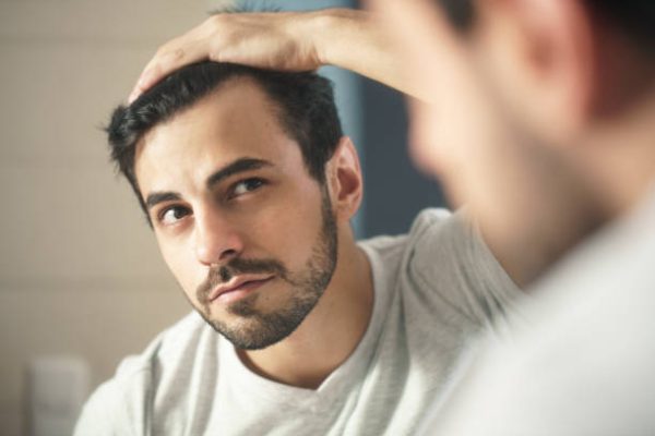 Latino person with beard grooming in bathroom at home. White metrosexual man worried for hair loss and looking at mirror his receding hairline.